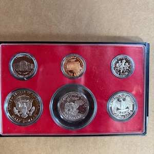 Op de afbeelding: Een set Amerikaanse munten, waaronder een halve dollar, een dollar, een kwart dollar, een dime en een penny, gepresenteerd in een zwarte behuizing met een rode binnenkant. De munten tonen verschillende ontwerpen en inscripties.