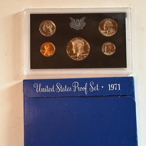 May include: A 1971 United States Proof Set in a clear plastic case, containing various coins. The set includes a half dollar, quarter, dime, nickel, and penny. The blue cardboard box below reads "United States Proof Set • 1971".