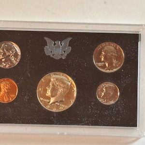 May include: A set of United States coins, including a penny, nickel, dime, quarter, and half-dollar, displayed in a protective case. The coins feature historical figures and symbols, with a silver eagle emblem at the top.