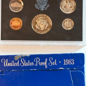 Könnte beinhalten: Ein 1983er United States Proof Set in einer Schutzhülle. Das Set enthält verschiedene Münzen, wie einen Penny, einen Dime und einen Half Dollar, alle mit detaillierten Designs. Die blaue Verpackung darunter trägt die Aufschrift "United States Proof Set - 1983".