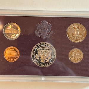 Könnte beinhalten: Ein Satz von US-Münzen, darunter ein Half Dollar, Quarter, Dime, Nickel und Penny, präsentiert in einer transparenten Schutzhülle. Die Münzen sind gold- und silberfarben und weisen detaillierte Designs und Inschriften auf.
