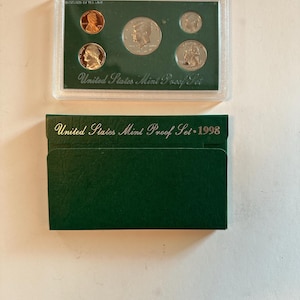 May include: A 1998 United States Mint Proof Set in a clear plastic case, containing several coins. The case sits atop a dark green cardboard sleeve with the text "United States Mint Proof Set • 1998".