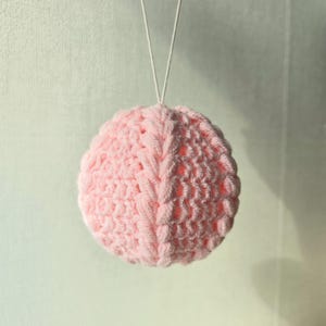 May include: A light pink, crocheted sphere ornament suspended by a white string. The sphere has a textured surface with a visible stitch pattern. The ornament is likely handmade and suitable for holiday or home decor.