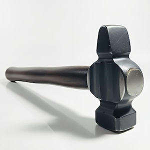 Puede incluir: Un martillo negro y plateado con un mango de madera oscura. La cabeza del martillo es de color gris oscuro con una cara de golpe plateada. El mango es de color marrón oscuro.