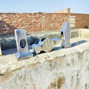 May include: Three silver metal hammers are displayed outdoors. The hammers are of different shapes and sizes, with one having a rectangular head with a circular hole. The other two hammers have a more traditional hammer shape.