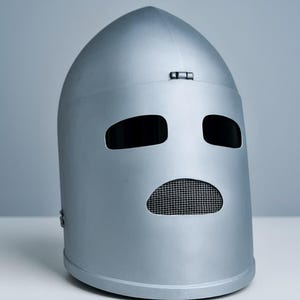 Puede incluir: Un casco plateado con una parte superior redondeada y un diseño similar a una cara. El casco tiene dos aberturas para los ojos y una abertura en forma de boca con una pantalla de malla. El casco es de metal liso.