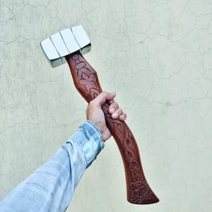 May include: A wooden hammer with a silver head and a brown handle. The handle is intricately carved with floral and geometric patterns. The hammer is held in a person's hand against a light-colored background.