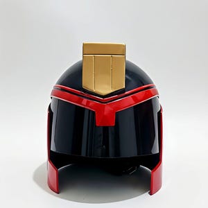 Könnte beinhalten: Schwarzer und roter Helm mit goldenem Akzent oben. Der Helm hat ein schlankes, futuristisches Design mit rotem Visier und Zierleisten. Die Oberseite weist ein goldenes rechteckiges Detail auf. Der Helm ist wahrscheinlich für Cosplay oder Kostüme gedacht.