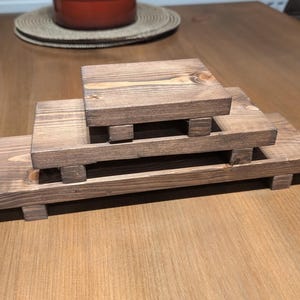 May include: A set of three wooden display risers in varying sizes, stained a warm brown. Each riser is rectangular with small, square feet. The risers are stacked, creating a tiered display for showcasing items.