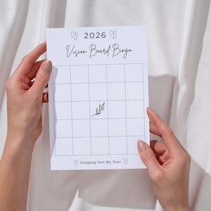 Può includere: Una scheda bianca di Vision Board Bingo per l'anno 2026, tenuta in mano. La scheda presenta una griglia, il titolo "Vision Board Bingo" e la frase "Stepping Into My Year". Sono presenti anche cuori decorativi e un rametto di foglie.