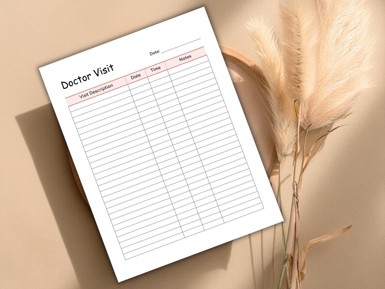 May include: A white Doctor Visit notepad with pink accents, featuring sections for visit description, date, time, and notes. The notepad is positioned on a neutral-toned surface, with dried ornamental grass in the background.