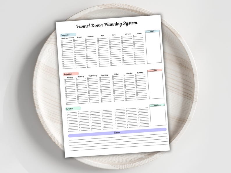 May include: A white planner sheet with the title "Funnel Down Planning System" in black font. The planner includes sections for categories, priorities, and schedules, with checkboxes and lines for notes. The planner is on a light wooden tray.