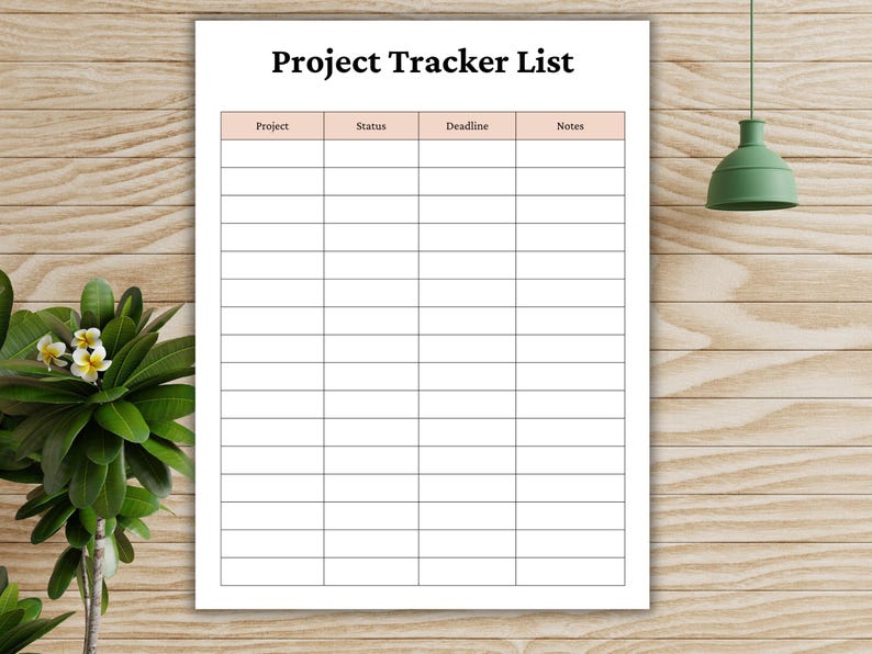 May include: A white Project Tracker List with a grid for tracking projects, status, deadlines, and notes. The title is in bold black font. The background is a light brown wood grain. A green lamp and plant are in the background.