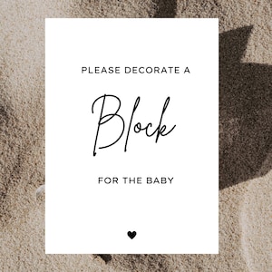 K&ouml;nnte beinhalten: Ein wei&szlig;es rechteckiges Schild mit schwarzem Text: &bdquo;BITTE DEKORIEREN SIE EINEN Block F&Uuml;R DAS BABY&ldquo; mit einem kleinen schwarzen Herz am unteren Rand. Das Schild befindet sich auf einer sandigen Oberfl&auml;che.