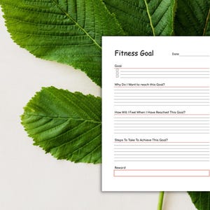May include: A white fitness goal planner with the title "Fitness Goal" and sections for goals, reasons, feelings, steps, and rewards. The planner is set against a backdrop of green leaves and a white surface.