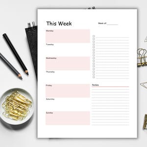 May include: A weekly planner with the title "This Week" and sections for each day, plus a notes section. Includes pencils, a small dish of gold paper clips, and binder clips. The planner has a white background with pink accents.