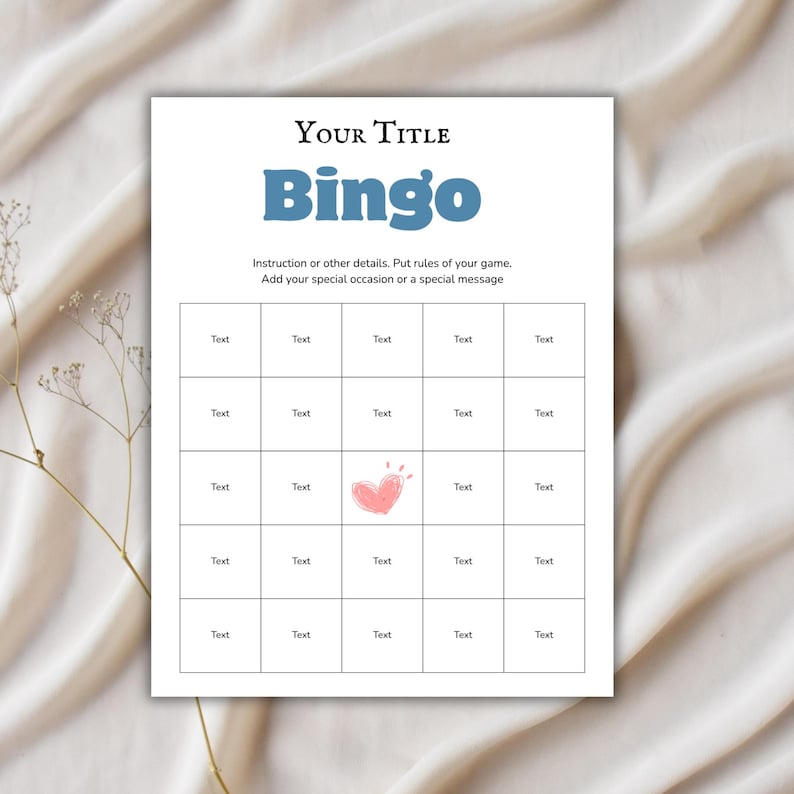 May include: A white Bingo game card with a grid of boxes, a pink heart graphic, and the word "Bingo" in blue. The card includes space for a title and instructions. The background is a light beige fabric.