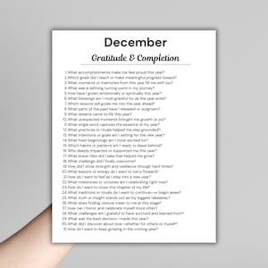 May include: A white paper with the title "December" and "Gratitude & Completion" printed in black. The paper contains a list of 31 questions for reflection, such as "What accomplishments make me feel proud this year?"