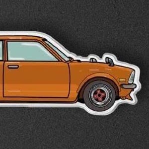 May include: Orange coupe car keyring with a white outline. The car has a two-door design with teal-coloured windows and black tyres with red accents. The keyring is on a dark grey background.