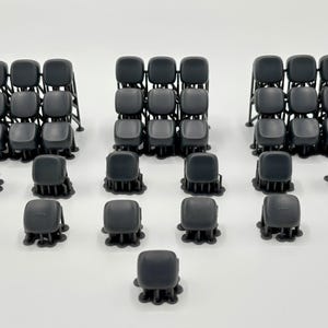 May include: A collection of dark gray, square-shaped resin keycaps arranged in three distinct grid-like clusters, accompanied by several individual loose keycaps, all featuring small support structures attached to their bases from the printing process.