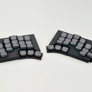 May include: Two black ergonomic split keyboards with gray keycaps. The keyboards are angled and designed to promote comfortable typing. The keycaps are a uniform gray color, and the overall design is modern and minimalist.