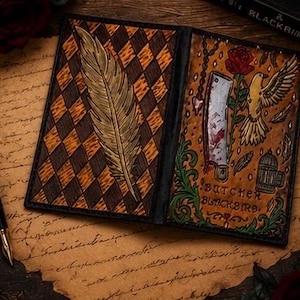 Puede incluir: Una cubierta de libro de cuero con un patrón de rombos en marrón claro y oscuro y una pluma grande a la izquierda. El lado derecho muestra una rosa roja, un cuchillo de carnicero, un pájaro y una jaula pequeña con el texto Butcher & Blackbird.