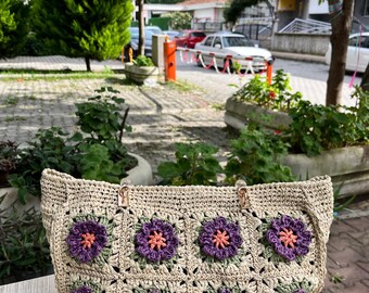 May include: A beige crochet tote bag with a floral pattern. The bag features purple and orange flower appliqués on a green background. The bag has two handles and a rectangular shape.