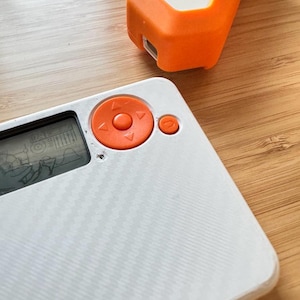 Pode incluir: Close-up de dois dispositivos eletrônicos. Um é branco com botões laranja e uma pequena tela exibindo uma imagem. O outro é laranja e branco. Ambos os dispositivos estão em uma superfície de madeira.