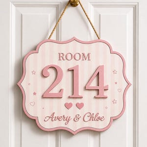 May include: A light pink and white striped wall sign with a scalloped edge, featuring the text ROOM 214 and the names Avery & Chloe. The sign includes raised pink numbers, two small pink hearts, and star icons, suspended by a tan rope.