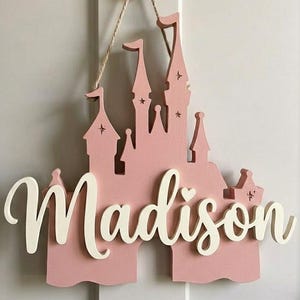 May include: A pink wooden castle-shaped sign with the name "Madison" in white script. The sign is hanging from a black hook on a white door. The castle has multiple towers and star cutouts.