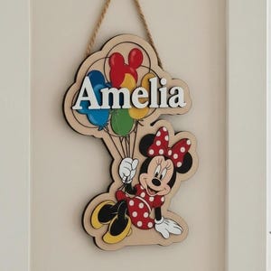May include: Wooden sign featuring Minnie Mouse holding balloons, with the name "Amelia" in white letters. The sign has a natural wood grain background and a black border, hanging from a rope. The balloons are red, blue, yellow, and green.