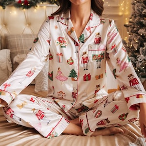May include: Cream-colored pajama set with a festive Christmas theme. The set features a button-down top and matching pants, both adorned with red trim and a repeating pattern of nutcrackers, ornaments, and wrapped gifts. The pajamas are made of a silky material.