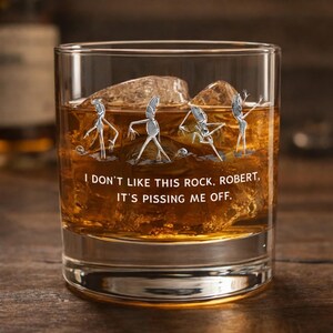 May include: A clear rocks glass containing amber liquid and ice cubes. The glass is decorated with a silver graphic of dancing figures and the text: "I DON'T LIKE THIS ROCK, ROBERT, IT'S PISSING ME OFF."