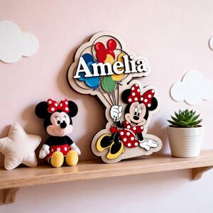 May include: A wooden wall decoration featuring Minnie Mouse holding balloons, with the name "Amelia" above. A Minnie Mouse soft toy and a star-shaped cushion are on a wooden shelf. The background is pale pink.