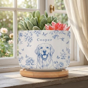 May include: White ceramic planter with a blue floral and dog design, the name "Cooper" is printed above the dog illustration. The planter holds green and red succulents and sits on a wooden saucer.