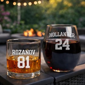 May include: Two personalized drinking glasses. One is a rocks glass with "ROZANOV 81" etched in white. The other is a stemless wine glass with "HOLLANDER 24" etched in white. The rocks glass contains amber liquid, and the wine glass contains red liquid.