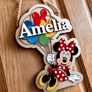 May include: Wooden sign featuring the name "Amelia" in white, with colorful balloons and a Minnie Mouse character. The sign is designed with a light brown background and a rope for hanging.
