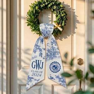 May include: A green wreath with a white and blue patterned sash. The sash has the text "GWJ CLASS OF 2024" and a university emblem. The wreath is hanging on a white door.