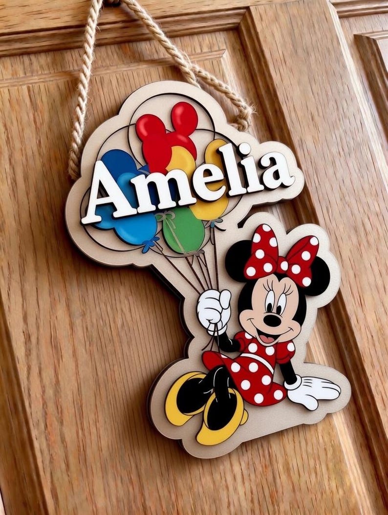 May include: Wooden sign with the name "Amelia" in white, accompanied by colourful balloons and a Minnie Mouse character. The sign is cloud-shaped and has a rope for hanging. Minnie Mouse is dressed in a red dress with white polka dots.