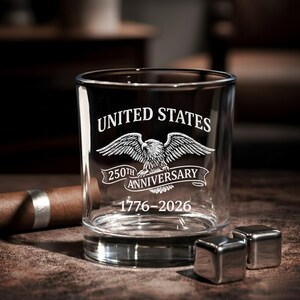 Może przedstawiać: Przezroczysty szklany kubek do whisky z białym napisem "UNITED STATES" i "250TH ANNIVERSARY" oraz grafiką orła. Daty "1776-2026" znajdują się poniżej. Dwie kostki lodu ze stali nierdzewnej znajdują się przed szklanką.