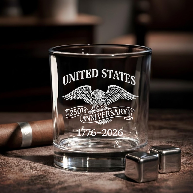 Może przedstawiać: Przezroczysty szklany kubek do whisky z białym napisem "UNITED STATES" i "250TH ANNIVERSARY" oraz grafiką orła. Daty "1776-2026" znajdują się poniżej. Dwie kostki lodu ze stali nierdzewnej znajdują się przed szklanką.