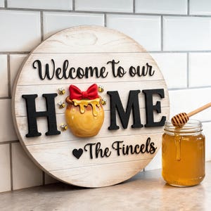 May include: A round wooden sign with a white faux-plank finish featuring the black text Welcome to our HOME and The Fincels. The letter O is replaced by a yellow honey pot with a red bow, surrounded by small bee illustrations and a black heart.