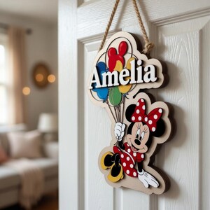 May include: Wooden door sign with Minnie Mouse holding balloons. The sign displays the name "Amelia" in white. Minnie Mouse is dressed in a red polka-dot dress and bow. The sign is suspended by a rope, suitable for a child's bedroom.