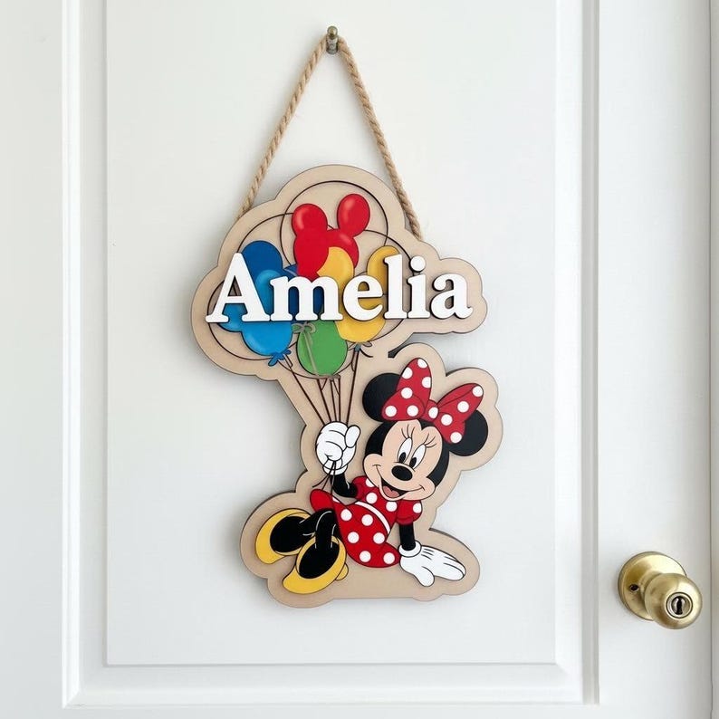 May include: Wooden door sign featuring the name "Amelia" in white letters, with a colorful Minnie Mouse design holding balloons. The sign is made of layered wood and hangs from a natural rope.