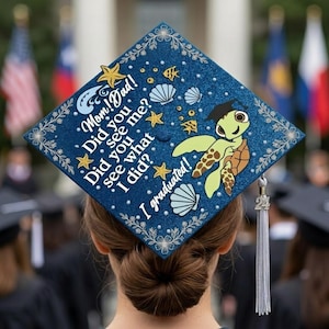Puede incluir: Un birrete azul marino decorado con una tortuga marina, conchas marinas y estrellas de mar. El birrete tiene el texto "Mom! Dad! Did you see me? Did you see what I did? I graduated!" y una borla plateada.