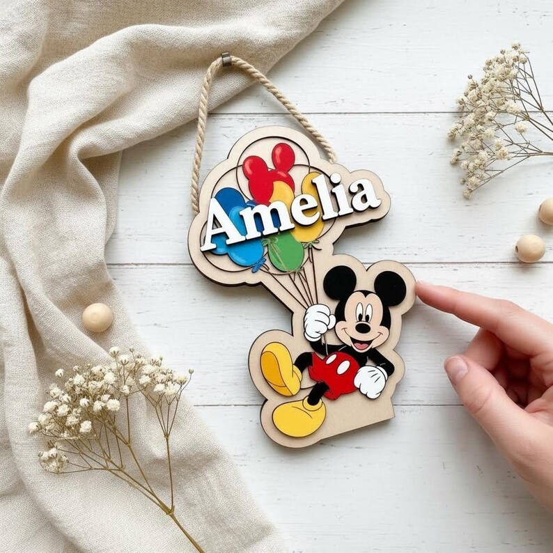 May include: Wooden sign featuring Mickey Mouse holding balloons with the name "Amelia" in white. The sign has a natural wood finish and a rope for hanging. Balloons are red, blue, yellow, and green.