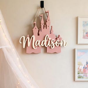 Pode incluir: Uma decora&ccedil;&atilde;o de parede em forma de castelo rosa com o nome "Madison" em fonte cursiva. O castelo &eacute; suspenso por uma corda de fio natural e montado numa parede de cor clara. Arte de parede decorativa para o quarto de uma crian&ccedil;a.