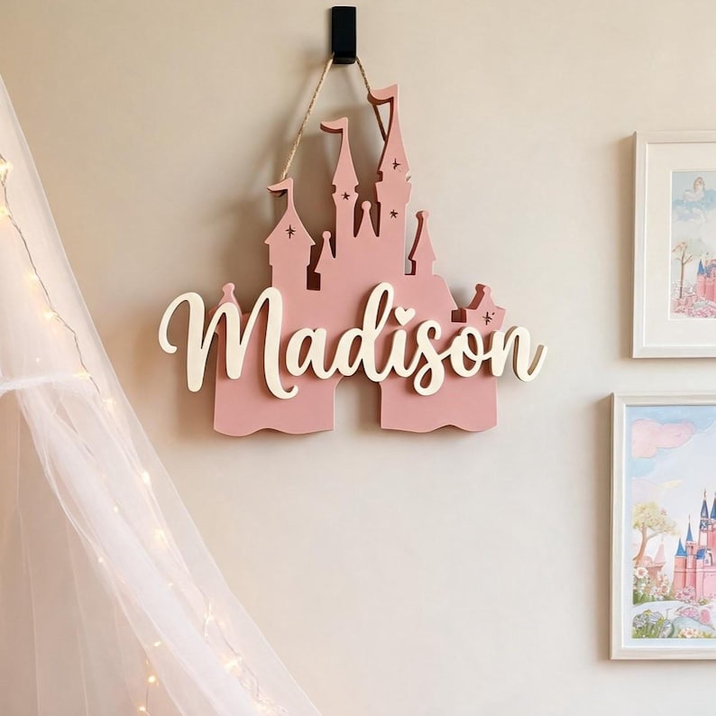 Pode incluir: Uma decora&ccedil;&atilde;o de parede em forma de castelo rosa com o nome "Madison" em fonte cursiva. O castelo &eacute; suspenso por uma corda de fio natural e montado numa parede de cor clara. Arte de parede decorativa para o quarto de uma crian&ccedil;a.
