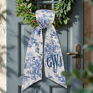 May include: A white and blue scarf with a toile pattern and the initials "GWJ" is draped over a blue door. The scarf is hanging on a door with a green wreath and a brass doorknob.