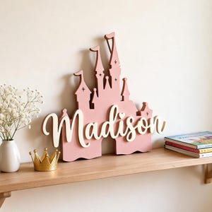 Pode incluir: Uma placa de madeira rosa em forma de castelo com o nome "Madison" em escrita branca. A placa &eacute; decorada com estrelas e fica em uma prateleira de madeira com uma coroa de ouro e livros.